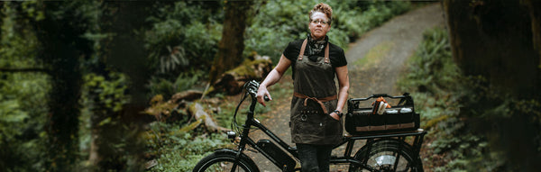 A woman stands in the woods with an electric cargo bike equipped with barbershop gear.