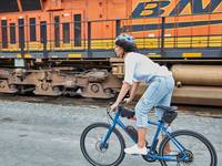 A woman of color wearing a helmet rides her blue RadMission high-step bike on a gravel road.