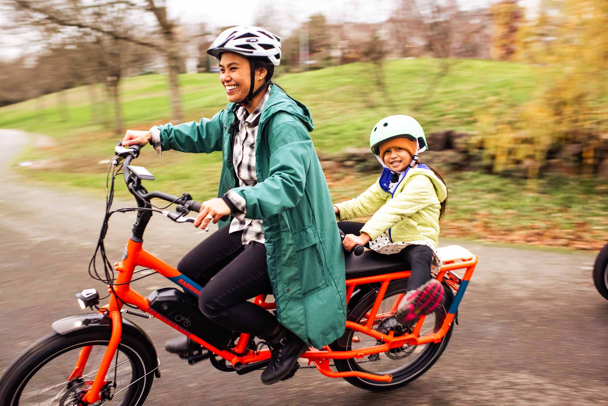 A mother and daughter ride a RadWagon through a park, with the daughter seated on the back smiling.