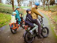 A woman of color rides an orange RadWagon with her daughter on the back.  A man rides a green RadRunner with a child on the back seat.